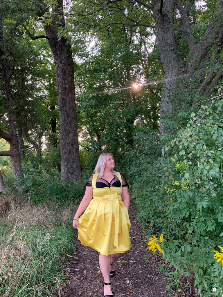 Confident woman wearing a bright yellow dirndl with black lace blouse, walking through a lush green forest at sunset.
